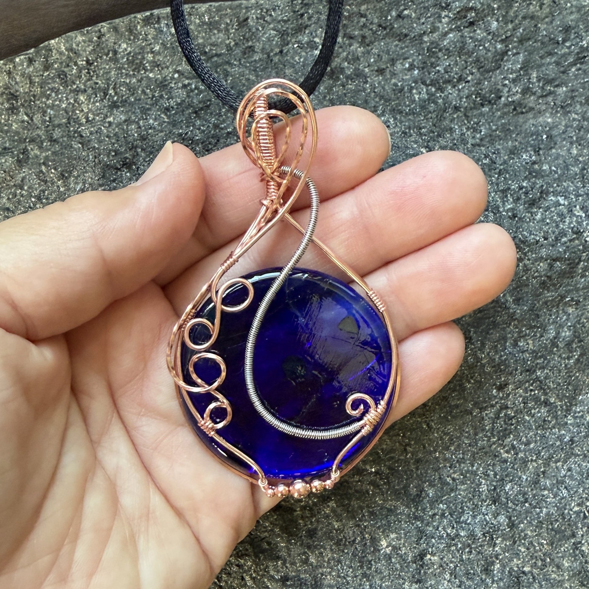 Front view of a teardrop-shaped, cobalt blue glass pendant necklace wrapped in copper and stainless steel wire, hanging from a black cord , held in the artist's hand.
