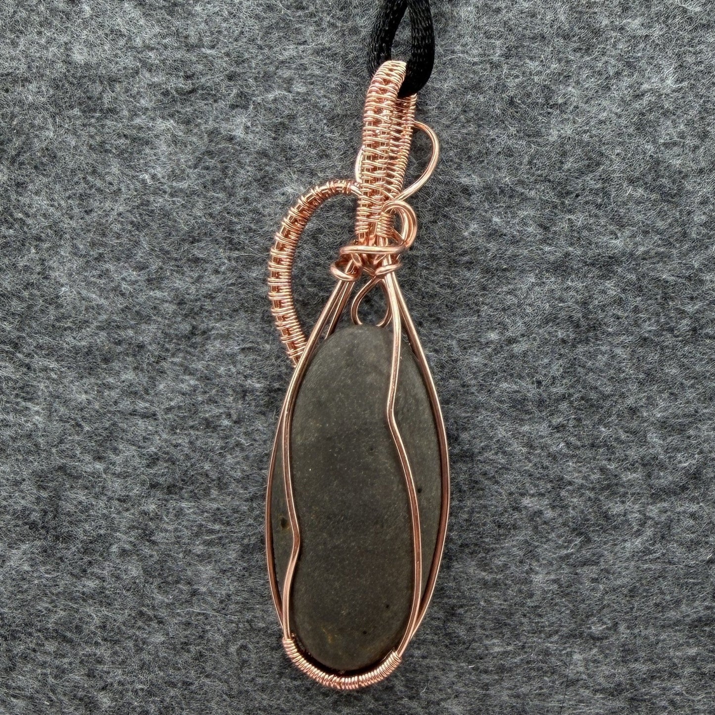 Back view of a narrow teardrop-shaped pendant necklace featuring a dark gray stone wrapped in copper wire, suspended from a black cord against a textured gray felt background.