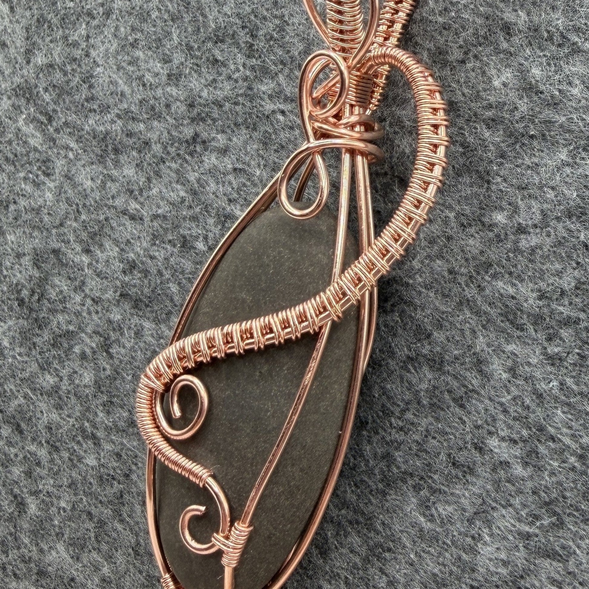 Detail view of a narrow teardrop-shaped pendant necklace featuring a dark gray stone wrapped in copper wire, suspended from a black cord against a textured gray felt background.