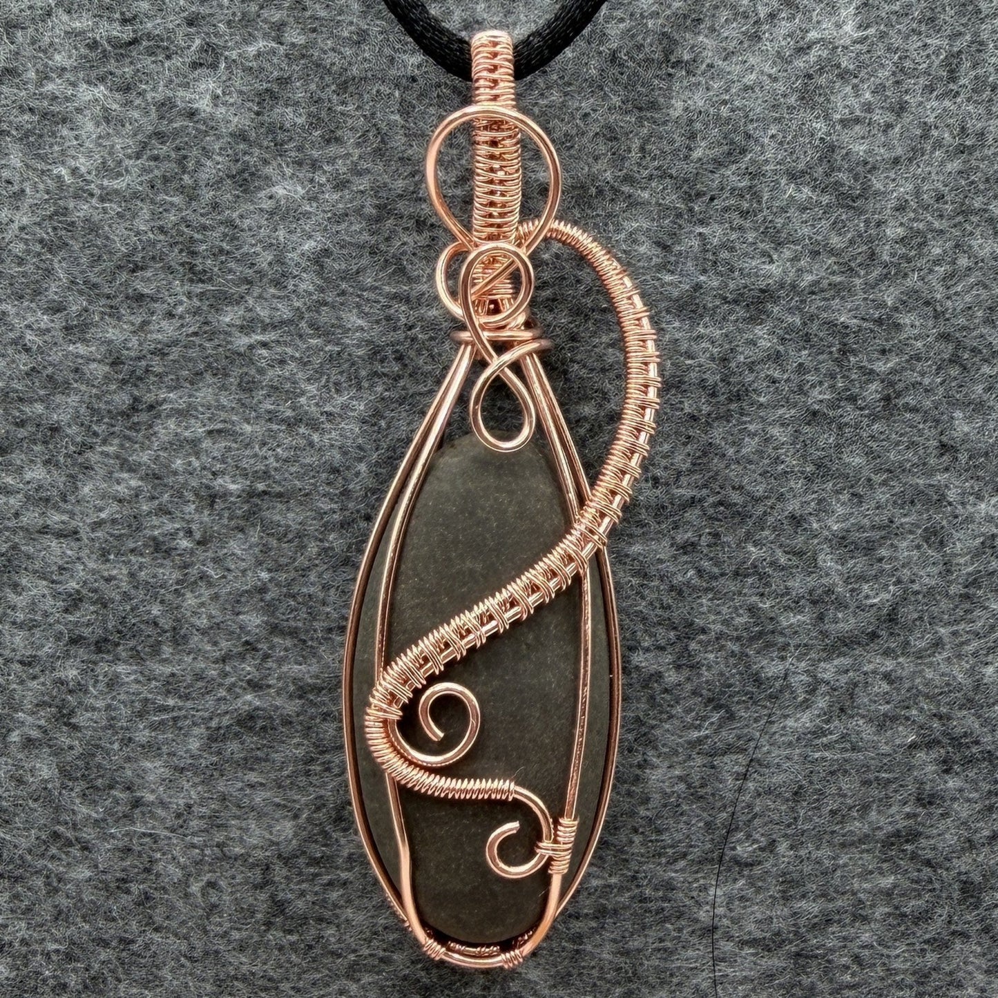 Front view of a narrow teardrop-shaped pendant necklace featuring a dark gray stone wrapped in copper wire, suspended from a black cord against a textured gray felt background.