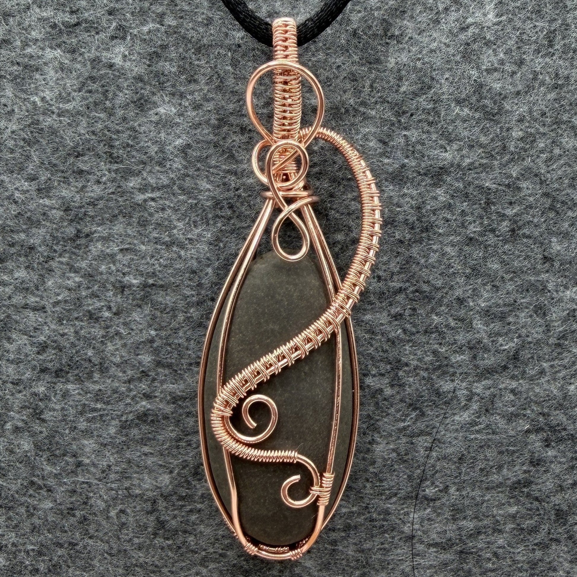 Front view of a narrow teardrop-shaped pendant necklace featuring a dark gray stone wrapped in copper wire, suspended from a black cord against a textured gray felt background.