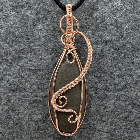 Front view of a narrow teardrop-shaped pendant necklace featuring a dark gray stone wrapped in copper wire, suspended from a black cord against a textured gray felt background.