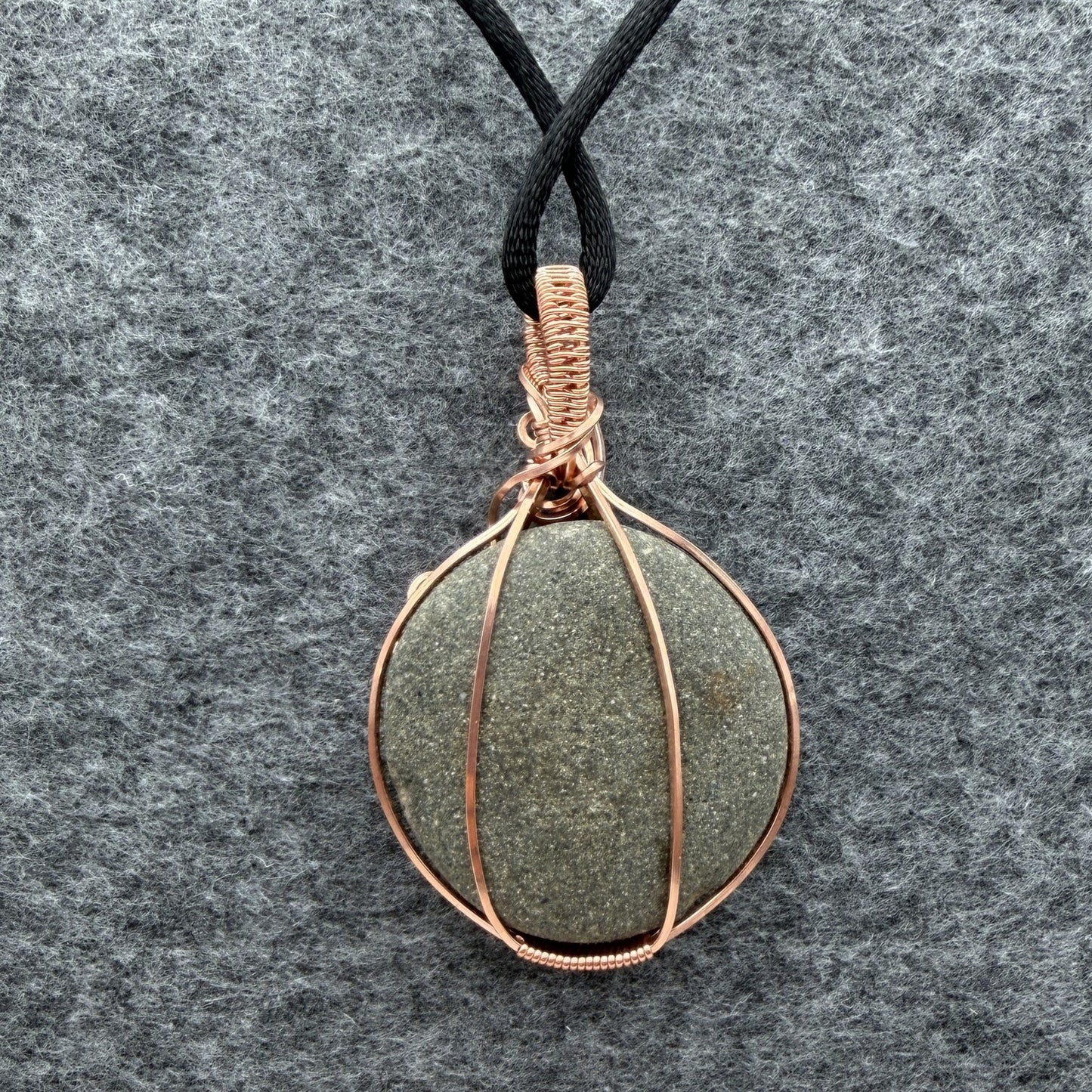 Back view of a handmade wire-wrapped pendant necklace featuring a round, dark gray stone with natural inclusions, wrapped in copper wire, hanging from a black cord on a textured gray background.