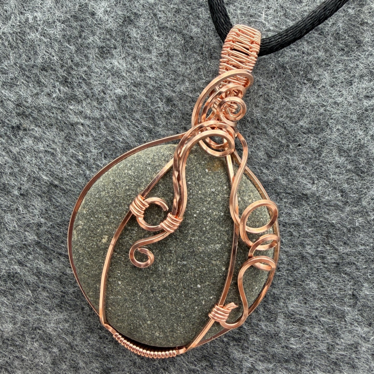 Detail view of a handmade wire-wrapped pendant necklace featuring a round, dark gray stone with natural inclusions, wrapped in copper wire, hanging from a black cord on a textured gray background.