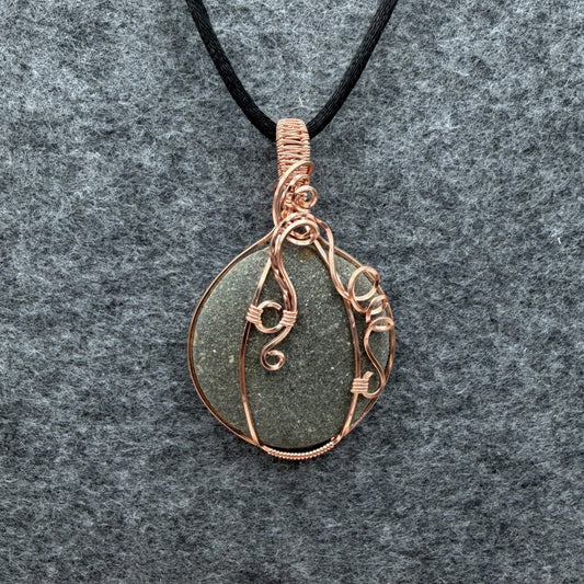 Front view of a handmade wire-wrapped pendant necklace featuring a round, dark gray stone with natural inclusions, wrapped in copper wire, hanging from a black cord on a textured gray background.