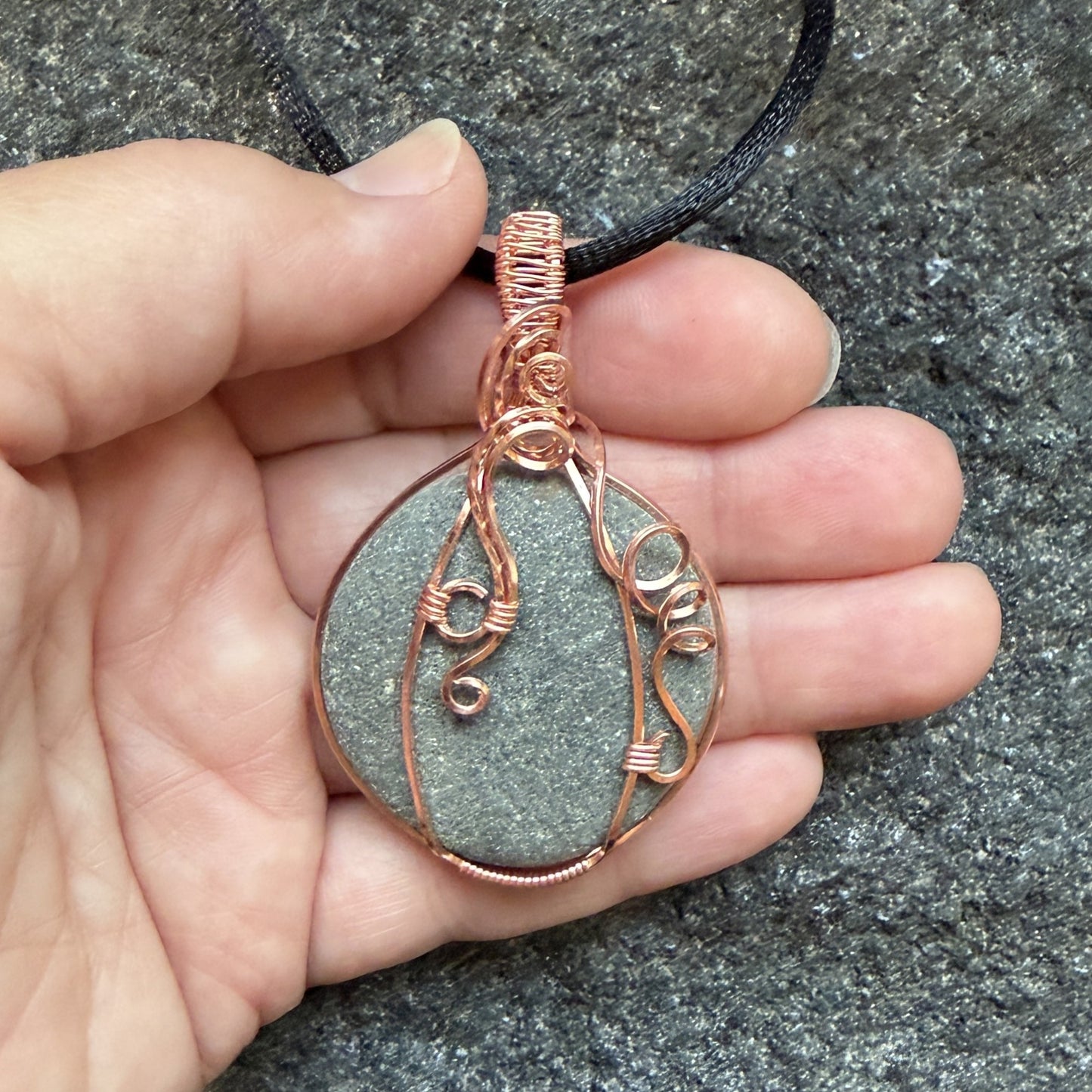 Front view of a handmade wire-wrapped pendant necklace featuring a round, dark gray stone with natural inclusions, wrapped in copper wire, hanging from a black cord, held in the artist's hand.
