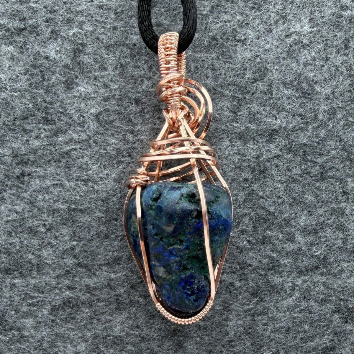 Back view of a handmade wire-wrapped pendant necklace featuring a blue and green stone with copper-colored wire wrapping, suspended from a black cord against a textured gray background.