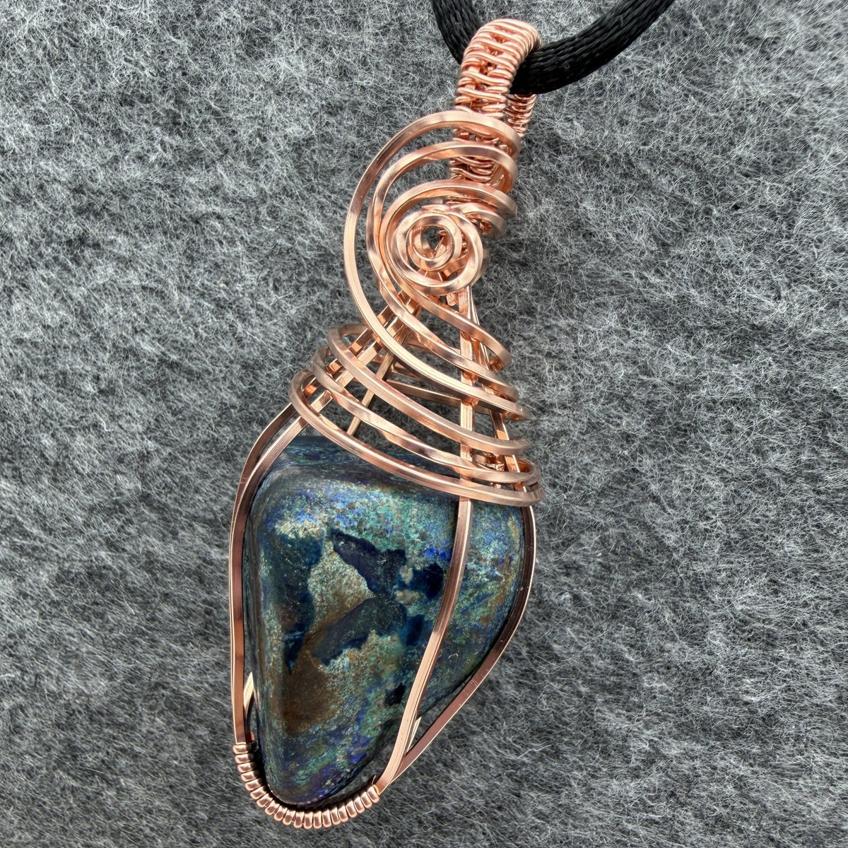Detail view of a handmade wire-wrapped pendant necklace featuring a blue and green stone with copper-colored wire wrapping, suspended from a black cord against a textured gray background.