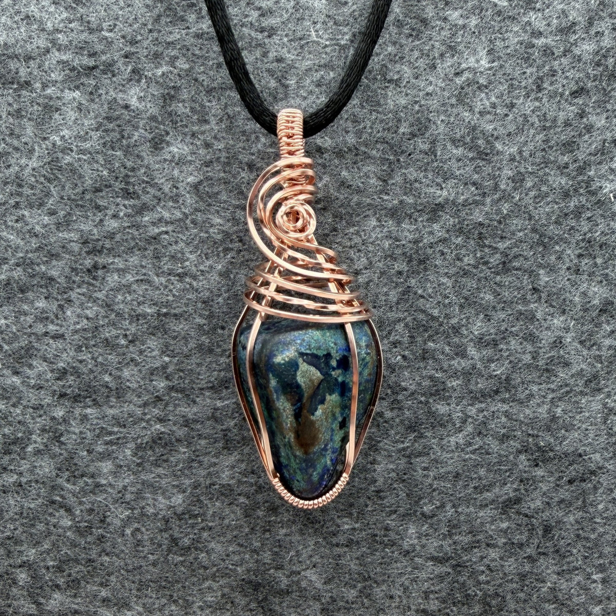 Front view of a handmade wire-wrapped pendant necklace featuring a blue and green stone with copper-colored wire wrapping, suspended from a black cord against a textured gray background.