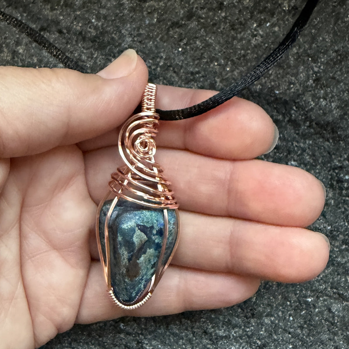 Front view of a handmade wire-wrapped pendant necklace featuring a blue and green stone with copper-colored wire wrapping, suspended from a black cord, held in the artist's hand.