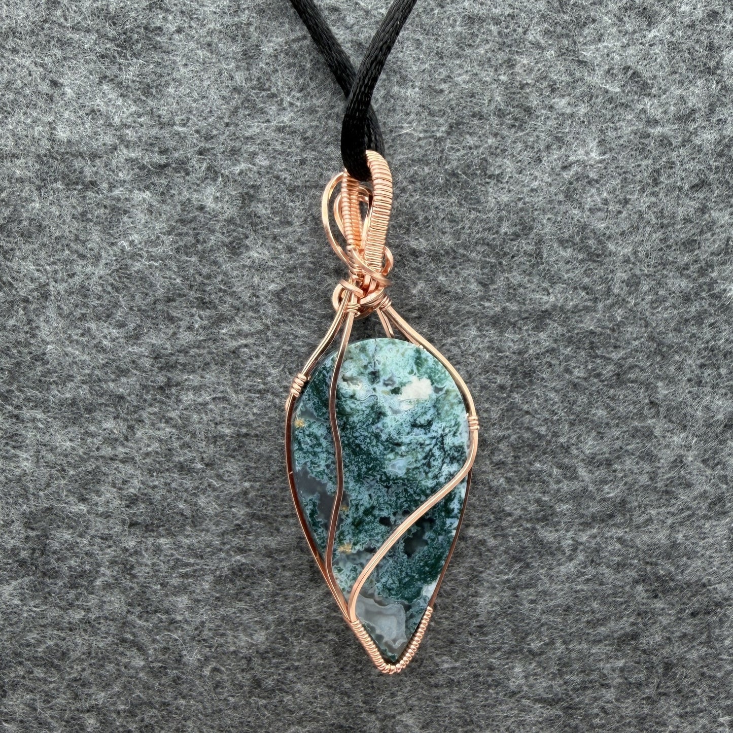 Back view of an inverted teardrop-shaped pendant necklace featuring a blue-green moss agate stone, wrapped in a copper wire frame and suspended from a black cord, set against a textured gray background.