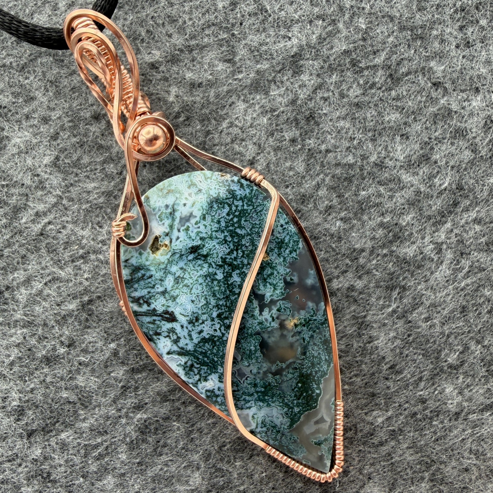 Detail view of an inverted teardrop-shaped pendant necklace featuring a blue-green moss agate stone, wrapped in a copper wire frame and suspended from a black cord, set against a textured gray background.