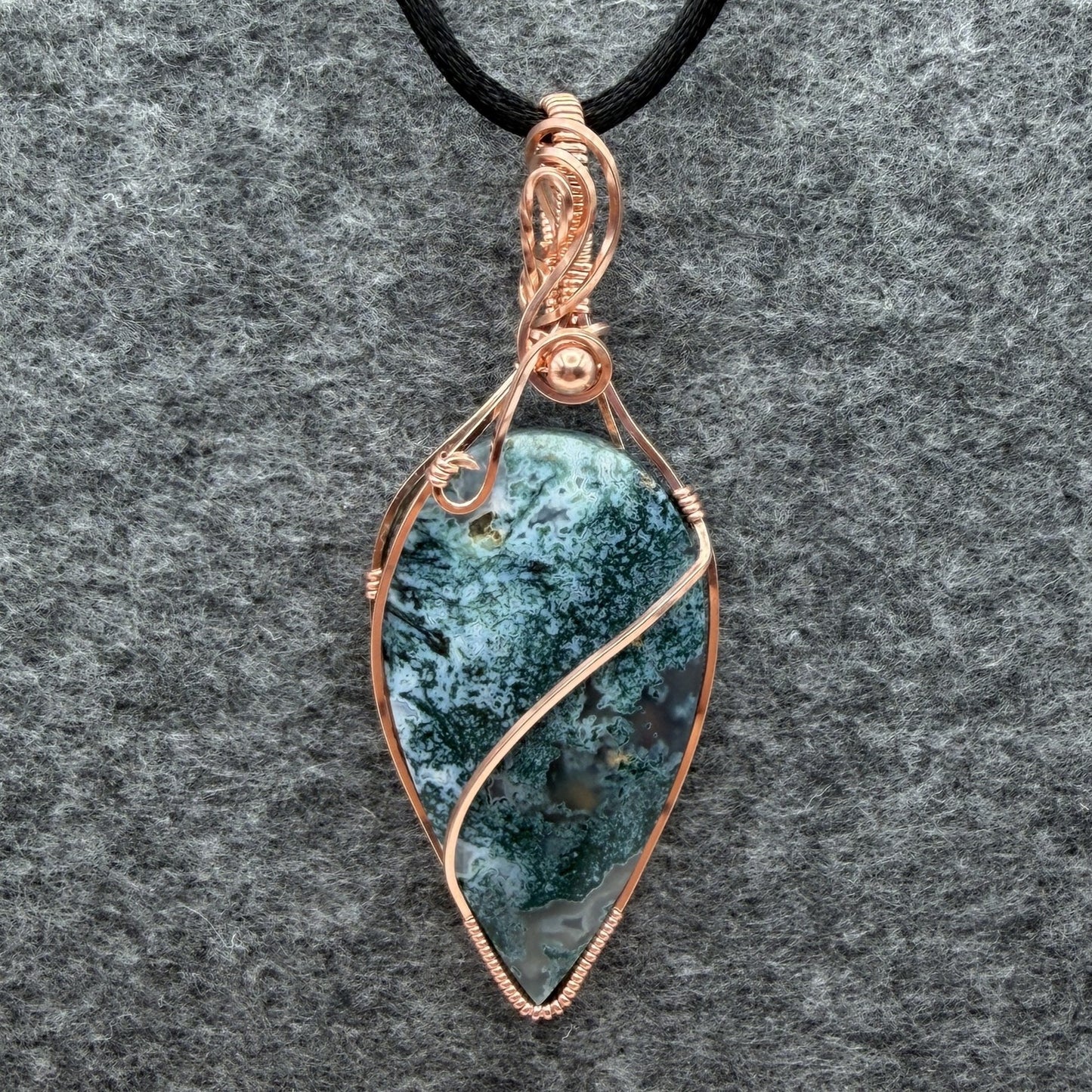 Front view of an inverted teardrop-shaped pendant necklace featuring a blue-green moss agate stone, wrapped in a copper wire frame and suspended from a black cord, set against a textured gray background.
