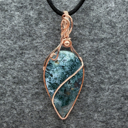 Front view of an inverted teardrop-shaped pendant necklace featuring a blue-green moss agate stone, wrapped in a copper wire frame and suspended from a black cord, set against a textured gray background.