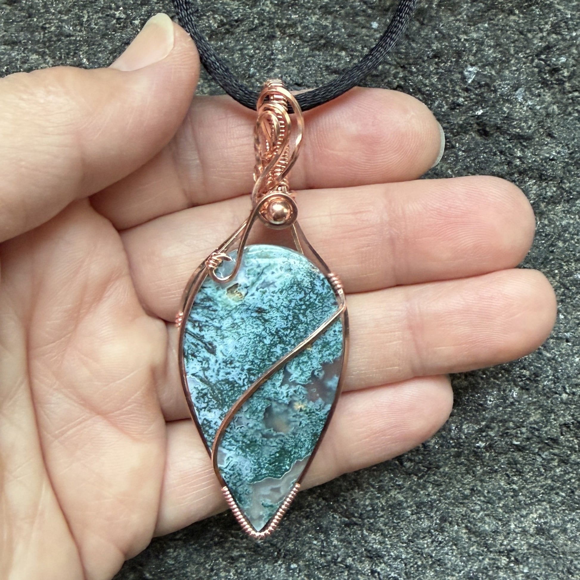 Front view of an inverted teardrop-shaped pendant necklace featuring a blue-green moss agate stone, wrapped in a copper wire frame and suspended from a black cord, held in the artist's hand.