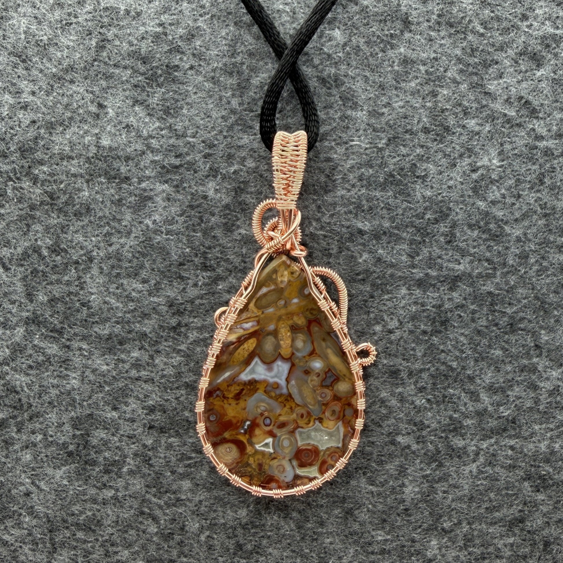 Back view of a teardrop-shaped pendant necklace featuring a brown and cream palm root agate stone wrapped in copper wire, suspended from a black cord against a textured gray background.