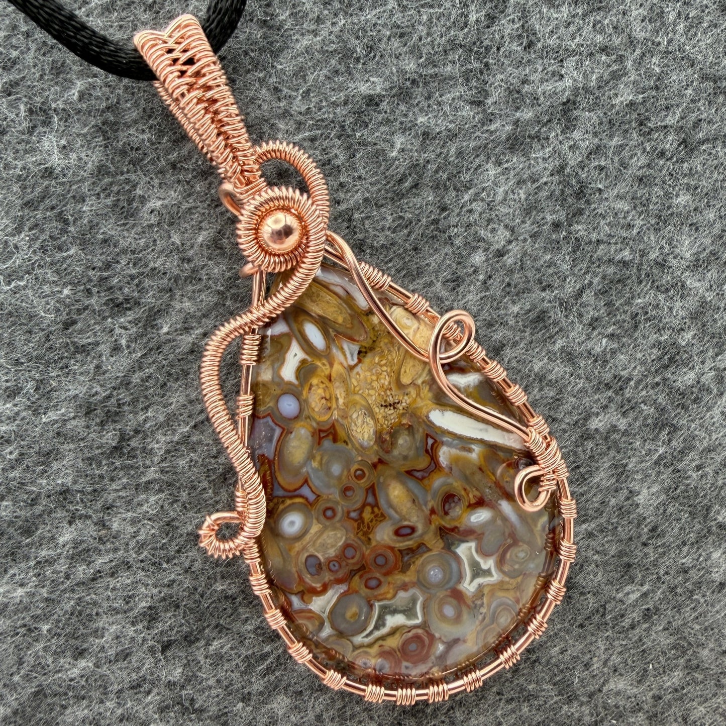 Detail view of a teardrop-shaped pendant necklace featuring a brown and cream palm root agate stone wrapped in copper wire, suspended from a black cord against a textured gray background.
