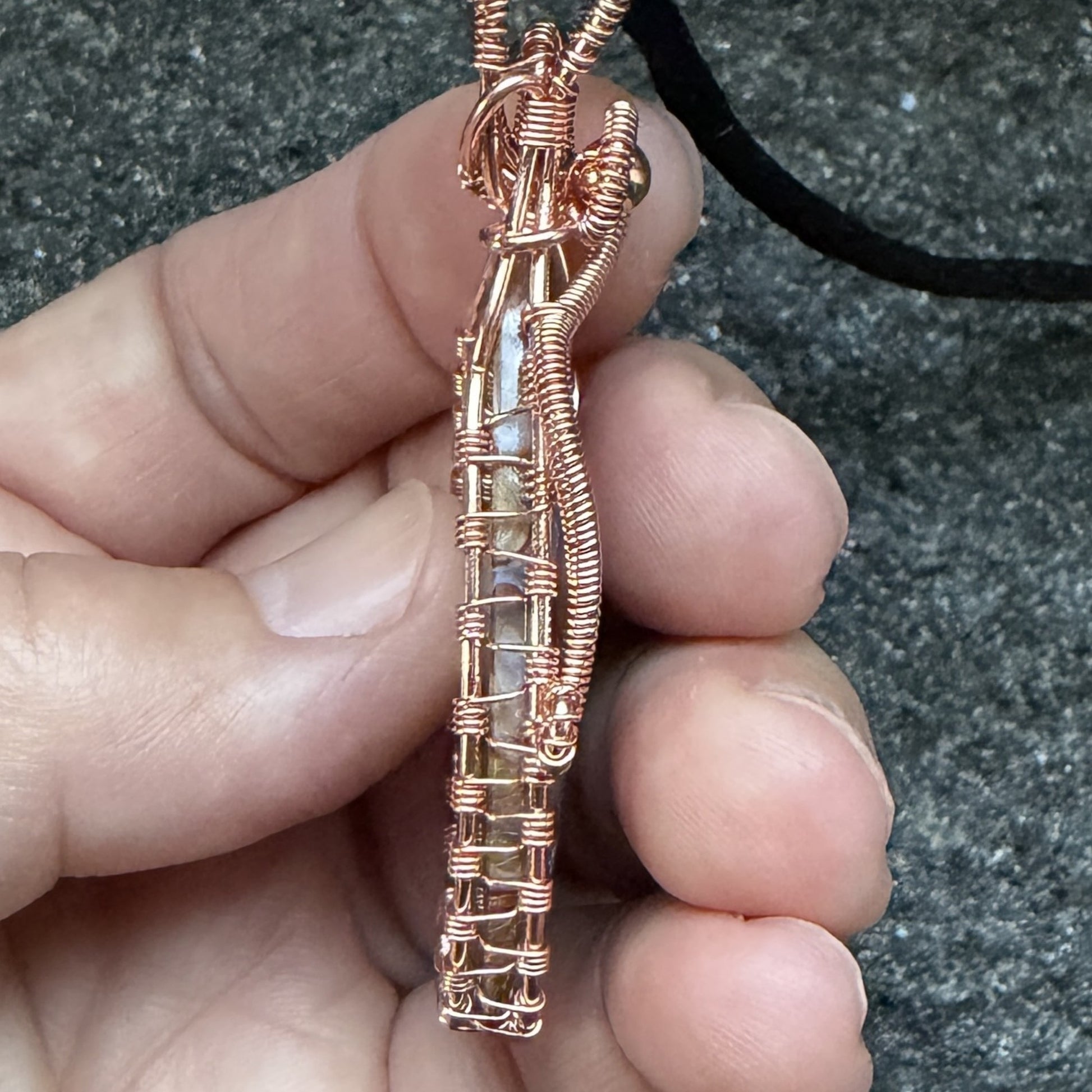 Detail view of the framing on a teardrop-shaped pendant necklace featuring a brown and cream palm root agate stone wrapped in copper wire, suspended from a black cord against a textured gray background.