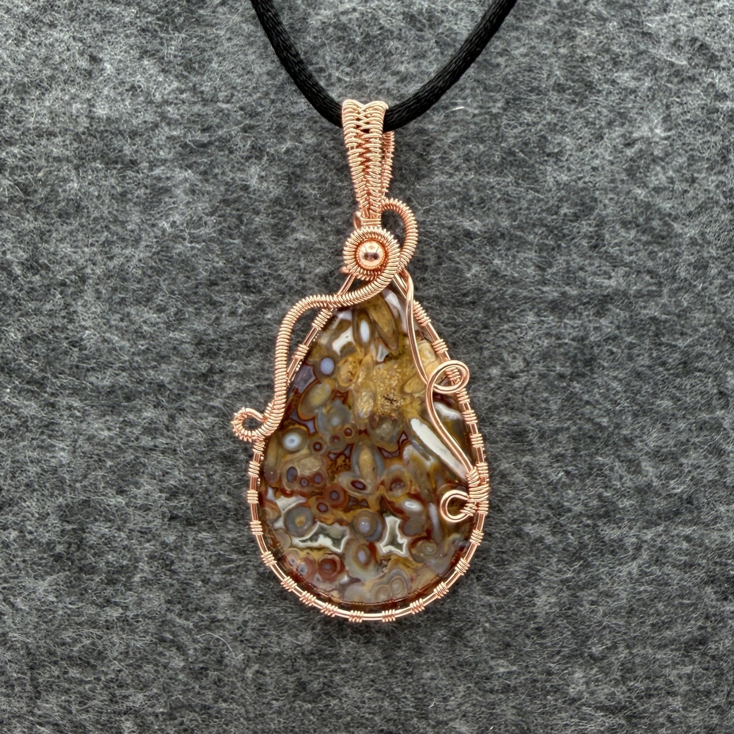 Front view of a teardrop-shaped pendant necklace featuring a brown and cream palm root agate stone wrapped in copper wire, suspended from a black cord against a textured gray background.