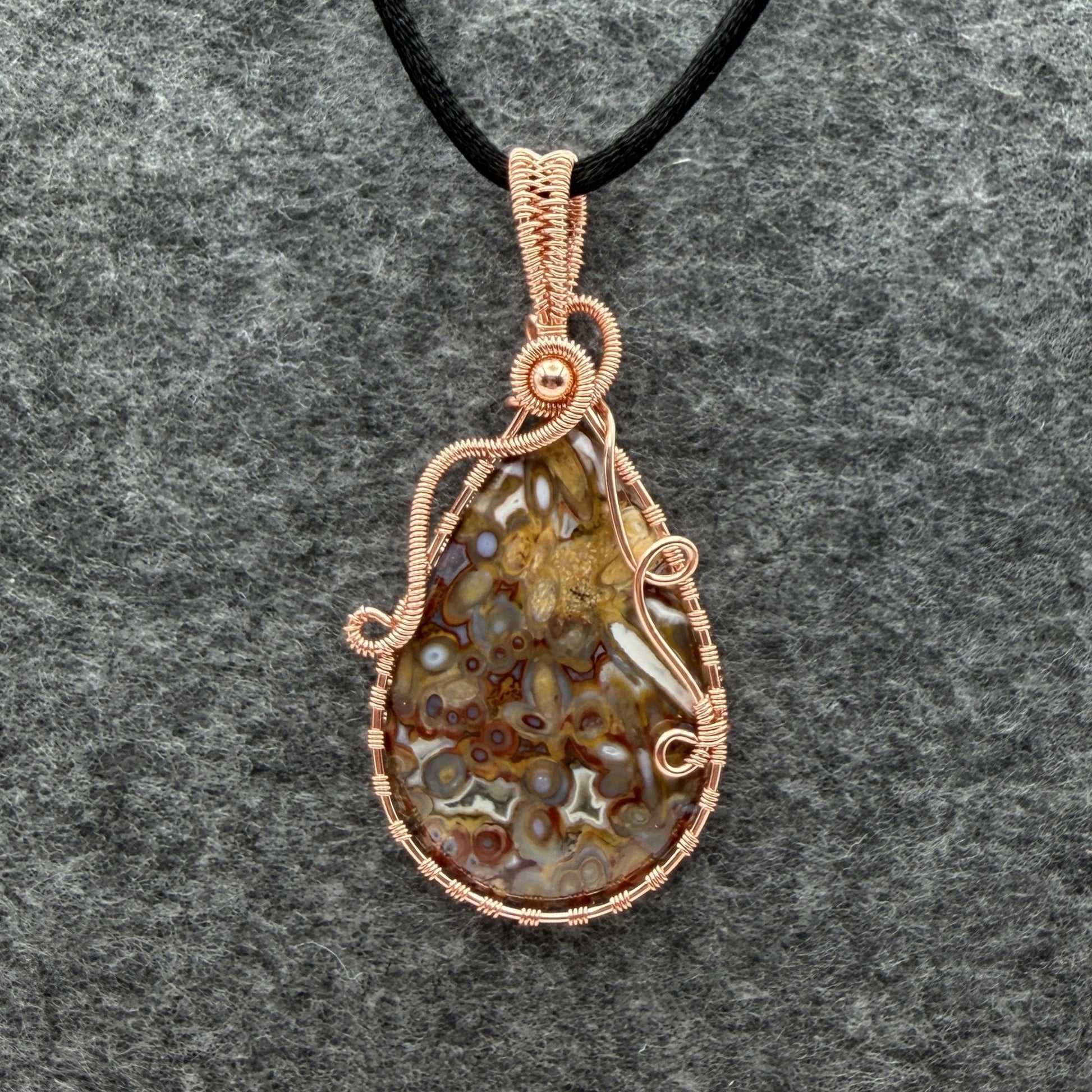Front view of a teardrop-shaped pendant necklace featuring a brown and cream palm root agate stone wrapped in copper wire, suspended from a black cord against a textured gray background.