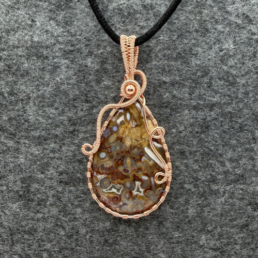 Front view of a teardrop-shaped pendant necklace featuring a brown and cream palm root agate stone wrapped in copper wire, suspended from a black cord against a textured gray background.
