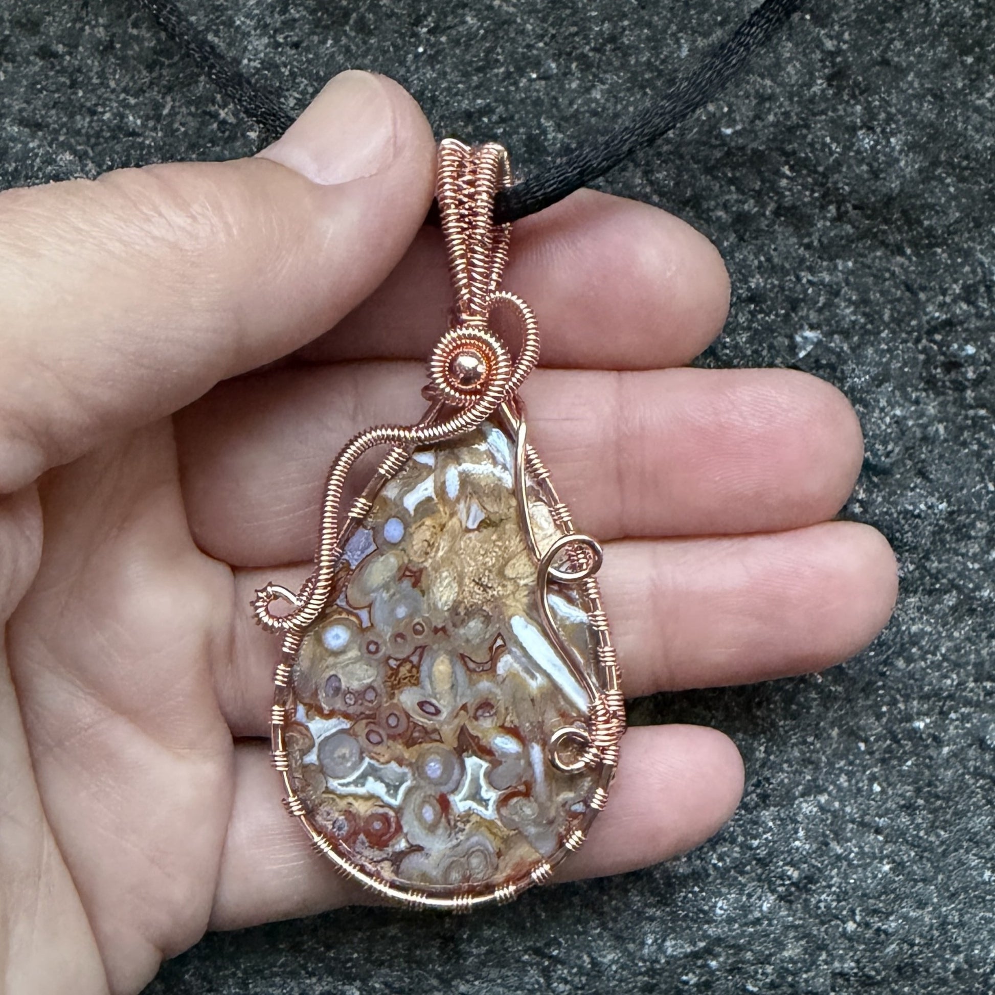 Front view of a teardrop-shaped pendant necklace featuring a brown and cream palm root agate stone wrapped in copper wire, suspended from a black cord, held in the artist's hand.