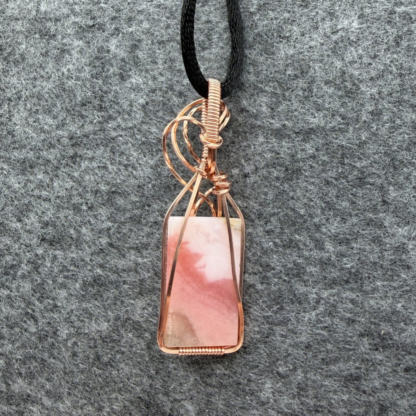 Back view of a layered blush pink stone and copper wire-wrapped pendant on a black cord, displayed on a textured gray felt background.