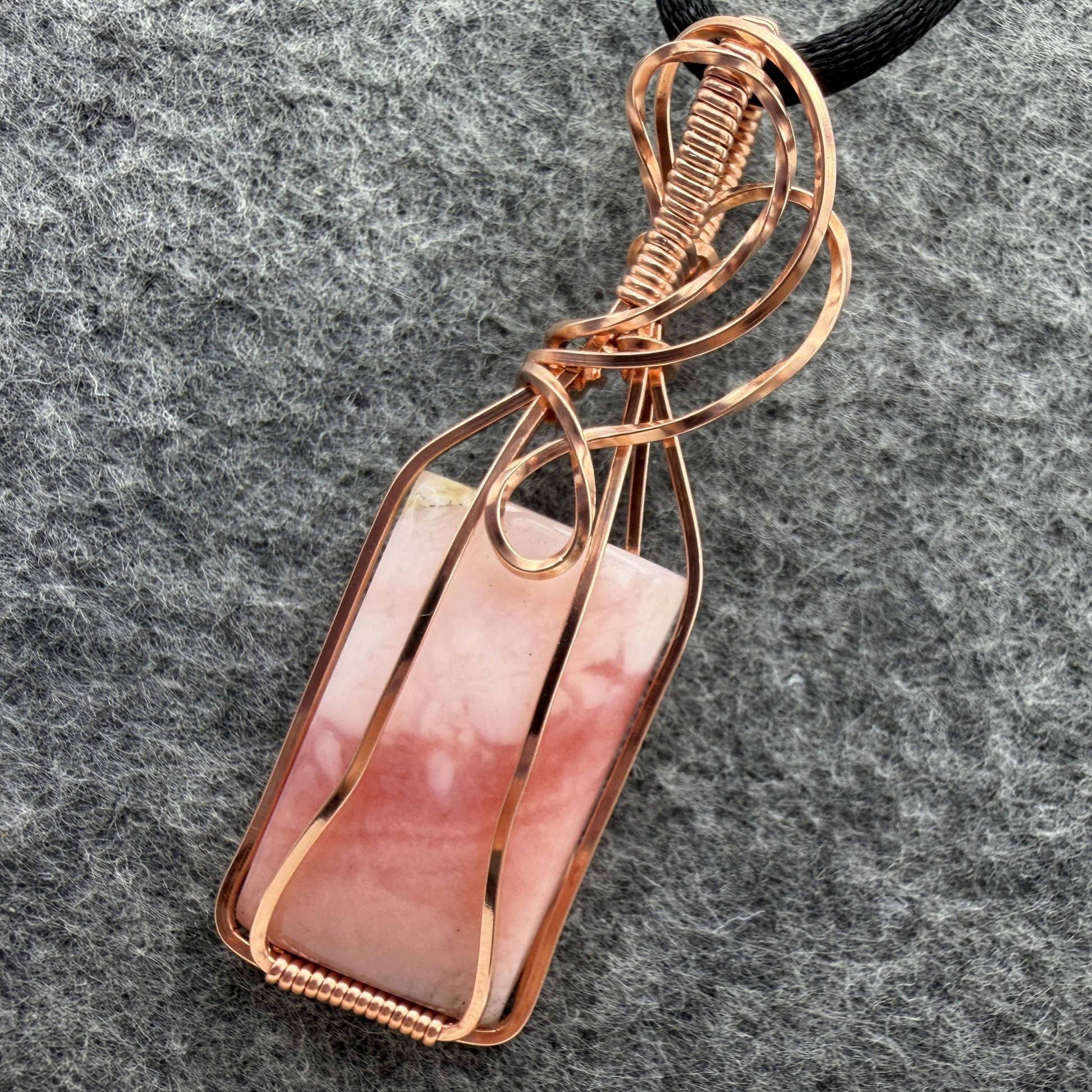 Close-up view of a layered blush pink stone and copper wire-wrapped pendant on a black cord, displayed on a textured gray felt background.
