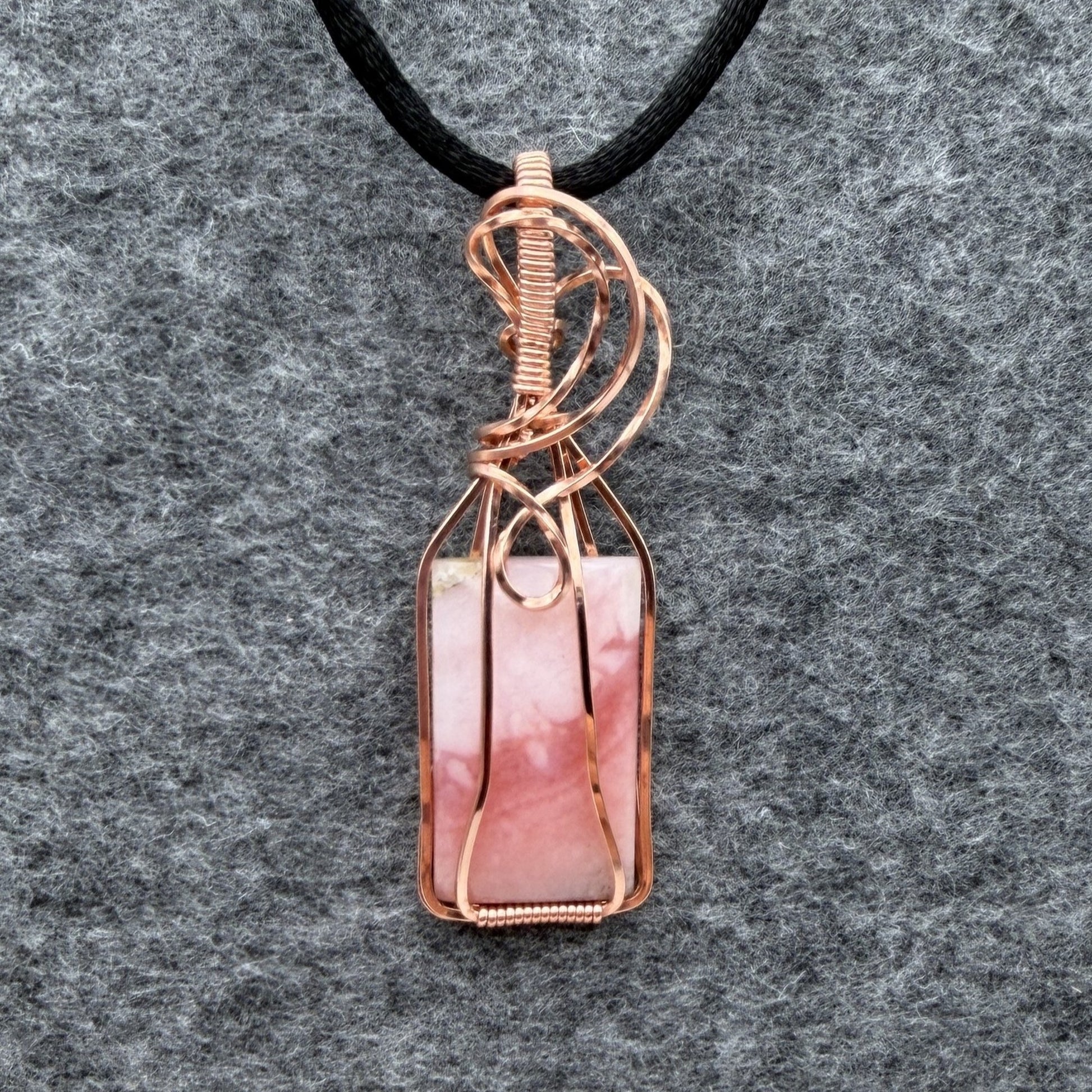 Front view of a layered blush pink stone and copper wire-wrapped pendant on a black cord, displayed on a textured gray felt background.
