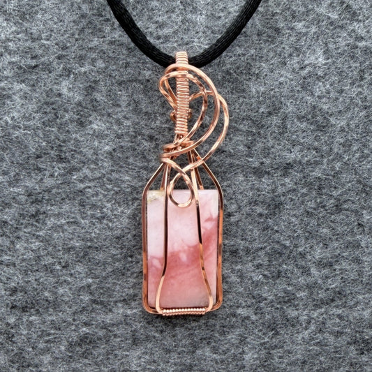 Front view of a layered blush pink stone and copper wire-wrapped pendant on a black cord, displayed on a textured gray felt background.