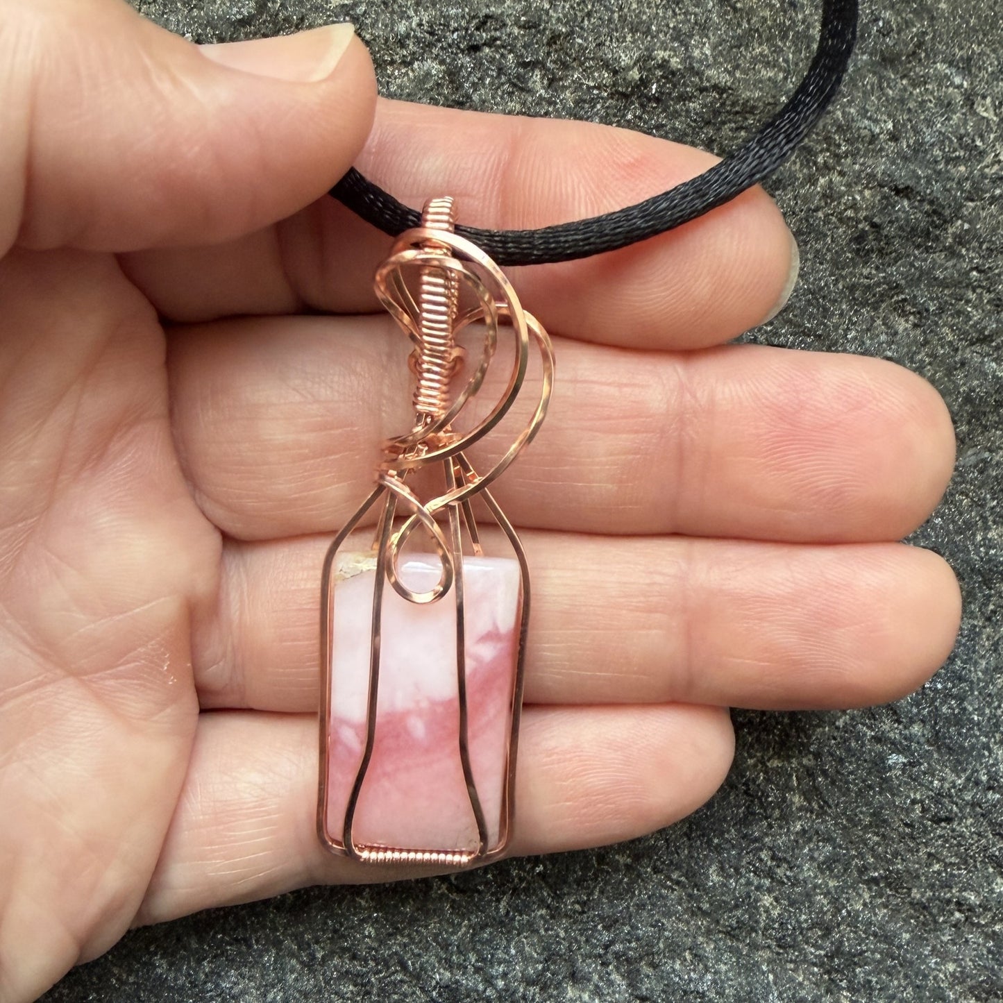 Front view of a layered blush pink stone and copper wire-wrapped pendant on a black cord, displayed in the artist's hand.
