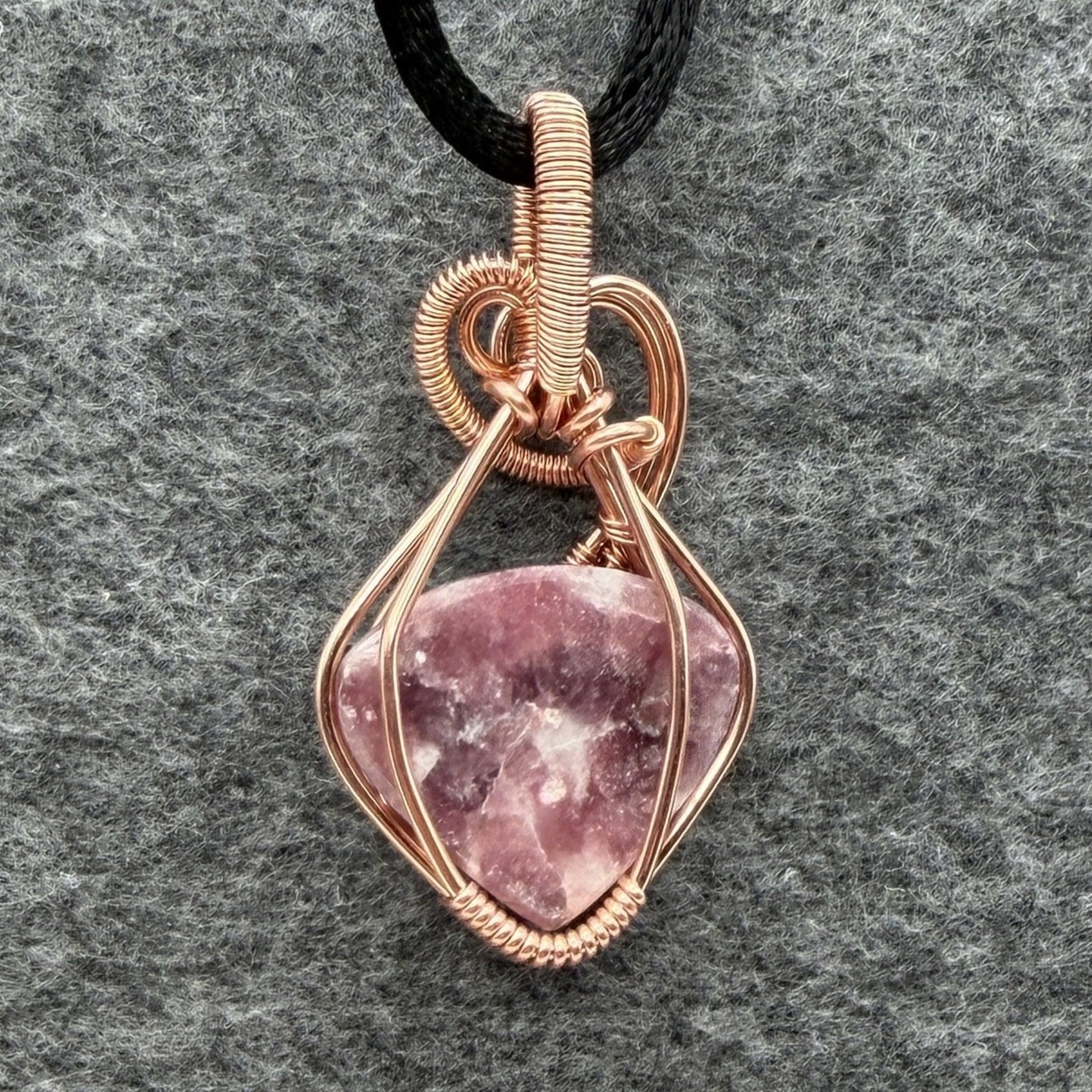 Back view of a copper wire-wrapped pendant necklace featuring a polished pinkish-purple quartz crystal, suspended from a black cord and displayed on a textured gray background.