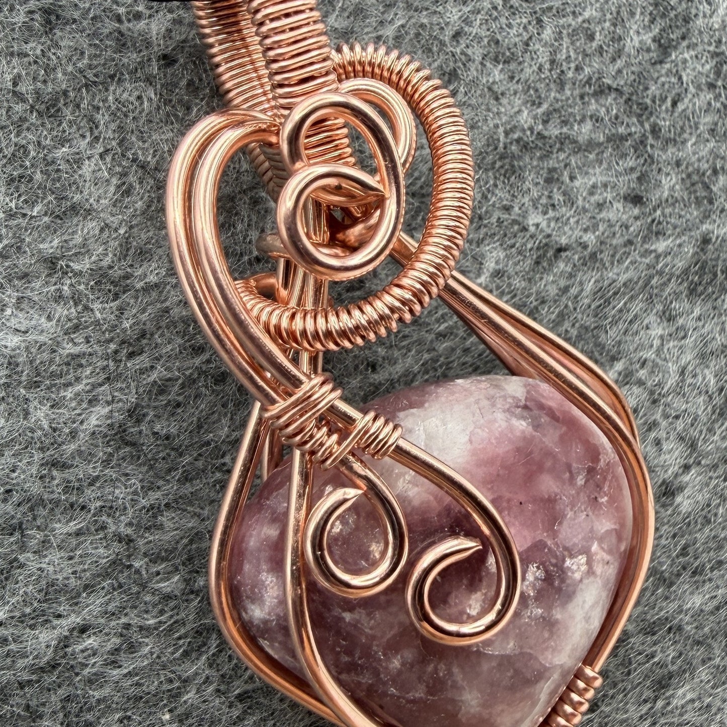 Detail view of a copper wire-wrapped pendant necklace featuring a polished pinkish-purple quartz crystal, suspended from a black cord and displayed on a textured gray background.