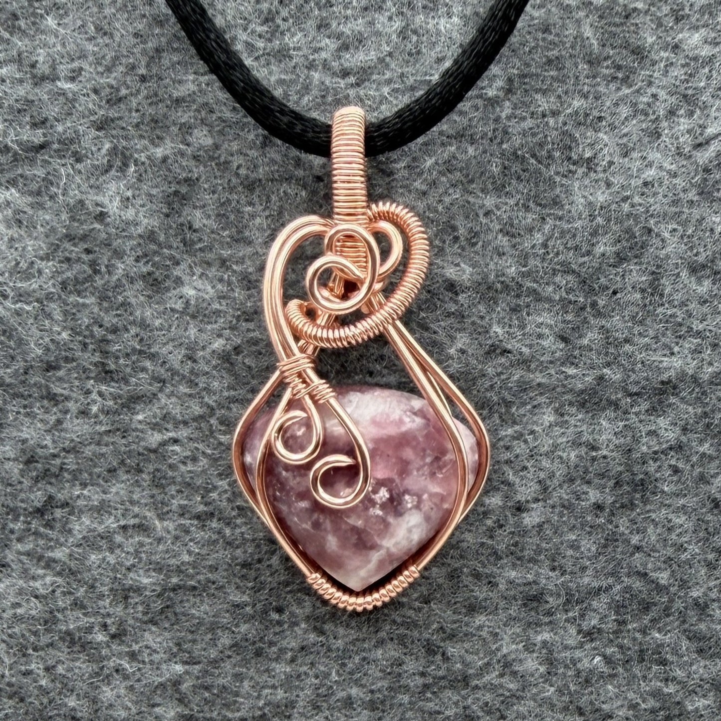Front view of a copper wire-wrapped pendant necklace featuring a polished pinkish-purple quartz crystal, suspended from a black cord and displayed on a textured gray background.