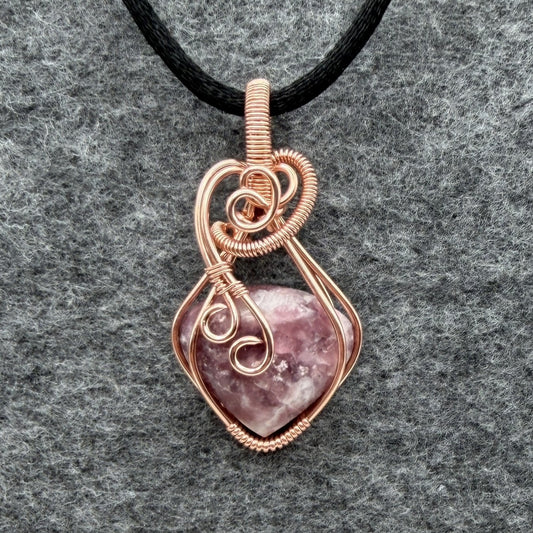 Front view of a copper wire-wrapped pendant necklace featuring a polished pinkish-purple quartz crystal, suspended from a black cord and displayed on a textured gray background.