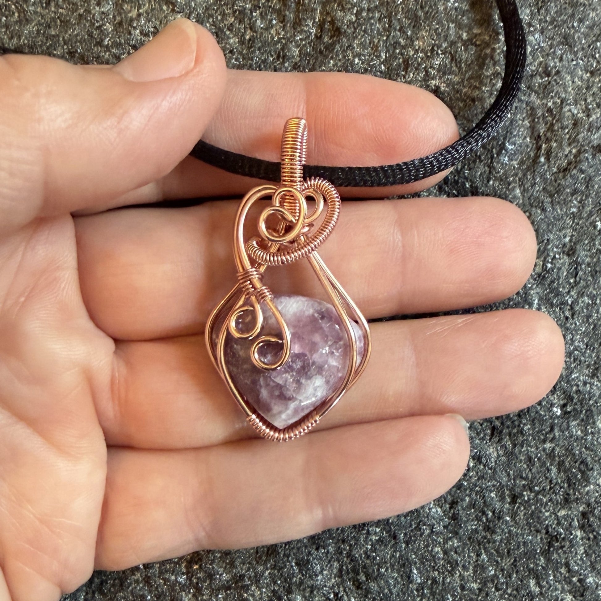 Front view of a copper wire-wrapped pendant necklace featuring a polished pinkish-purple quartz crystal, suspended from a black cord and held in the artist's hand.