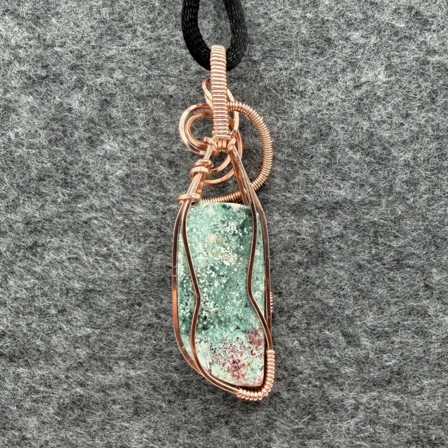 Back view of a wire-wrapped pendant necklace featuring a teal marbled stone with a cranberry-colored tip hanging from a black cord against a textured gray background.