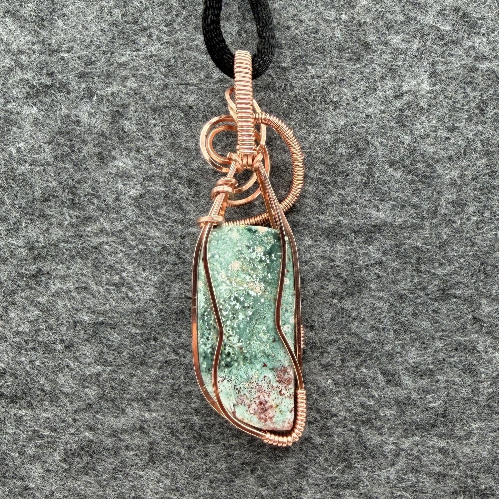 Back view of a wire-wrapped pendant necklace featuring a teal marbled stone with a cranberry-colored tip hanging from a black cord against a textured gray background.