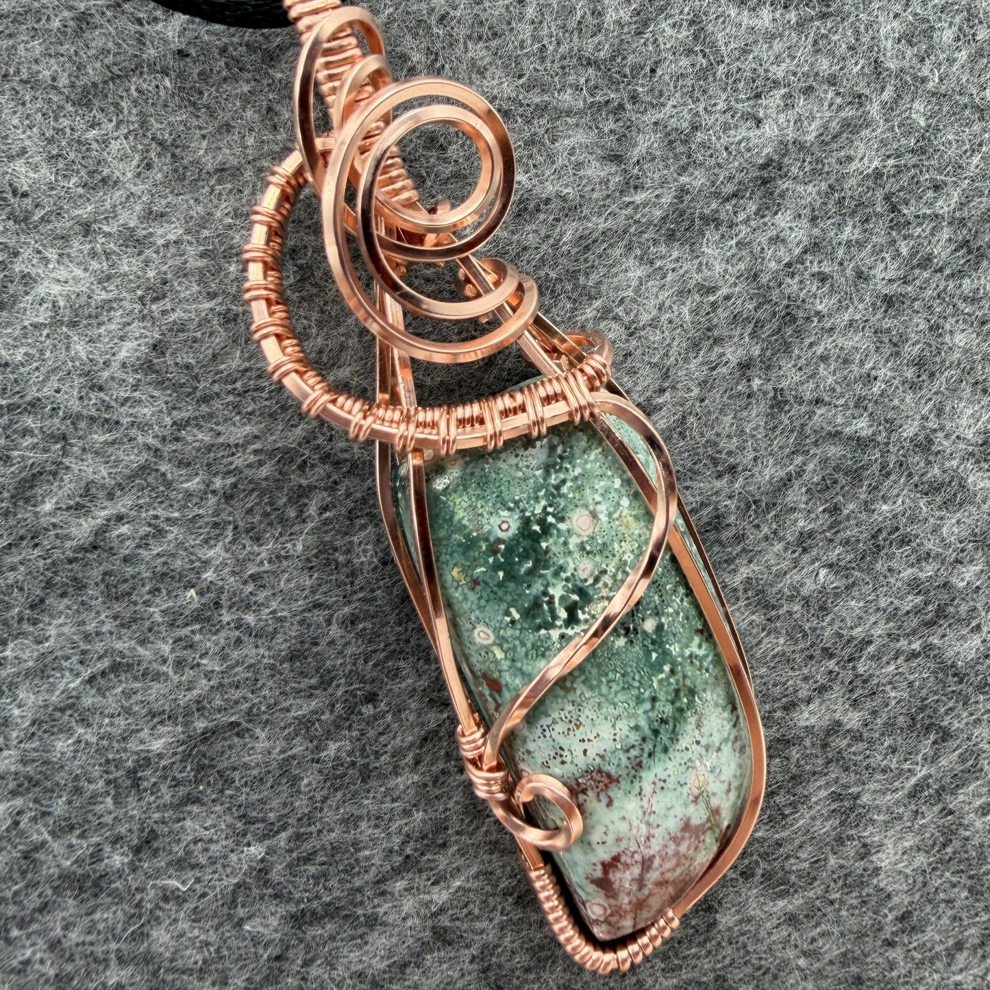 Detail view of a wire-wrapped pendant necklace featuring a teal marbled stone with a cranberry-colored tip hanging from a black cord against a textured gray background.