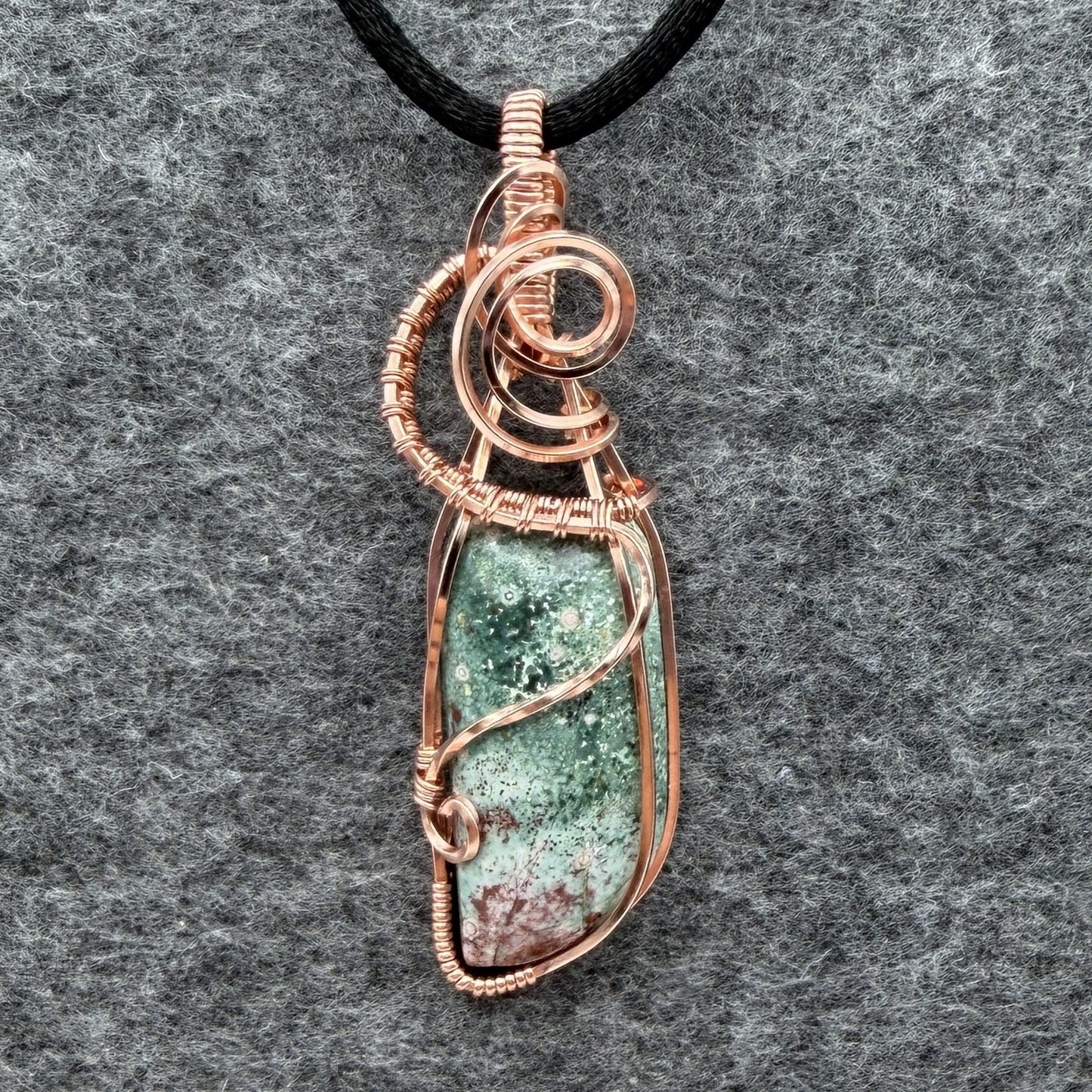 Front view of a wire-wrapped pendant necklace featuring a teal marbled stone with a cranberry-colored tip hanging from a black cord against a textured gray background.