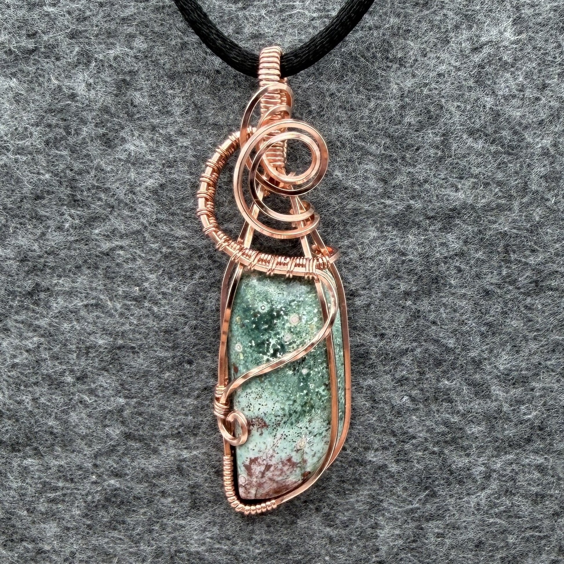 Front view of a wire-wrapped pendant necklace featuring a teal marbled stone with a cranberry-colored tip hanging from a black cord against a textured gray background.