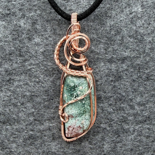 Front view of a wire-wrapped pendant necklace featuring a teal marbled stone with a cranberry-colored tip hanging from a black cord against a textured gray background.