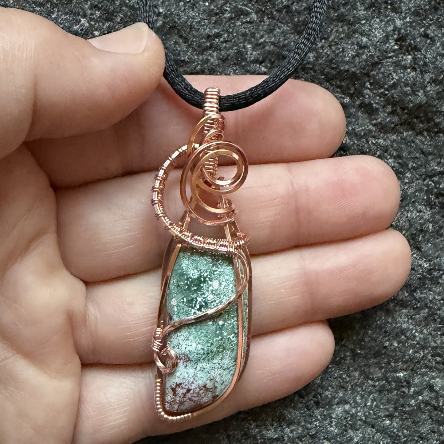Front view of a wire-wrapped pendant necklace featuring a teal marbled stone with a cranberry-colored tip hanging from a black cord held in the artist's hand.