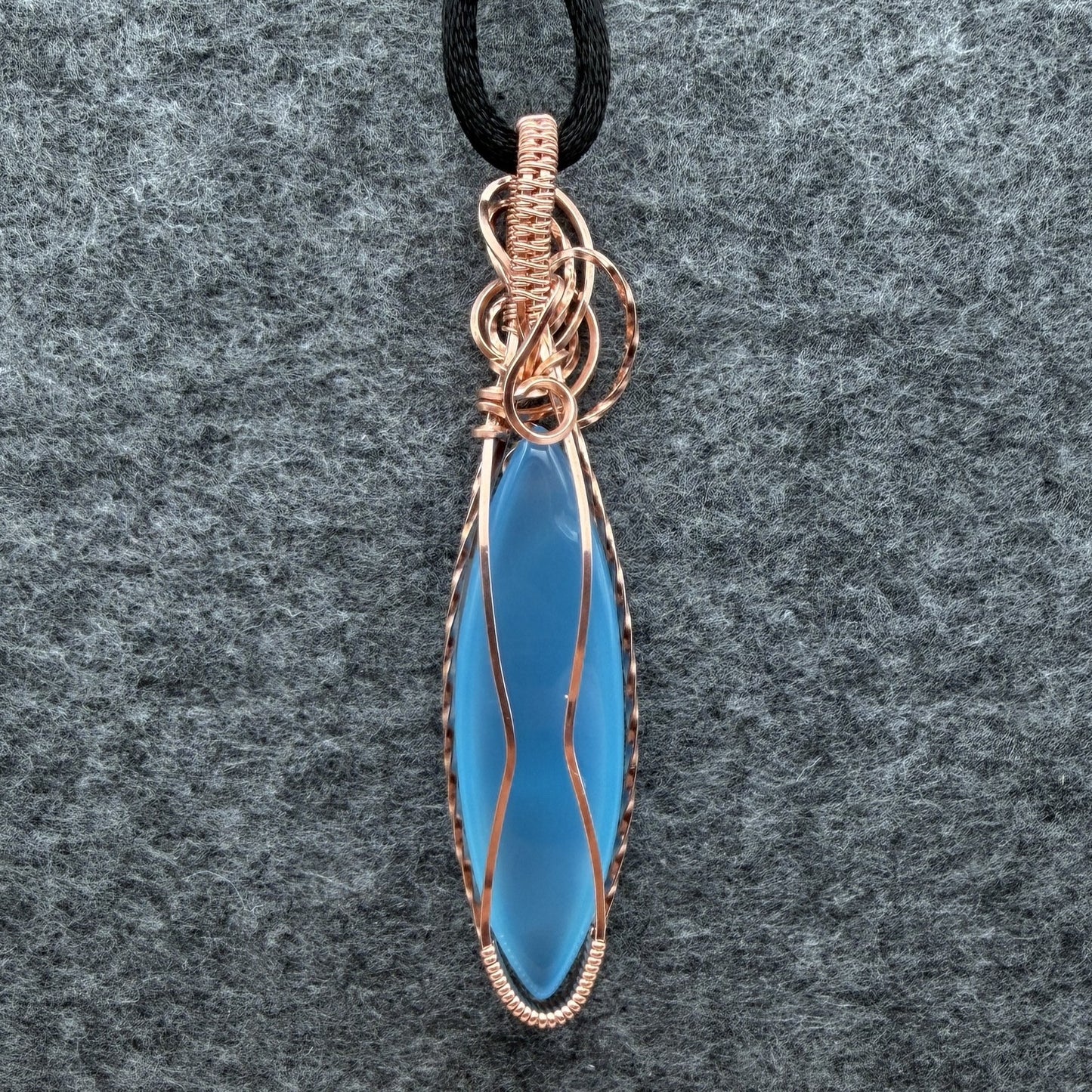 Back view of a handmade pendant featuring a smooth, marquise-shaped light blue stone wrapped in textured copper wire, hanging from a black cord against a gray felt background.
