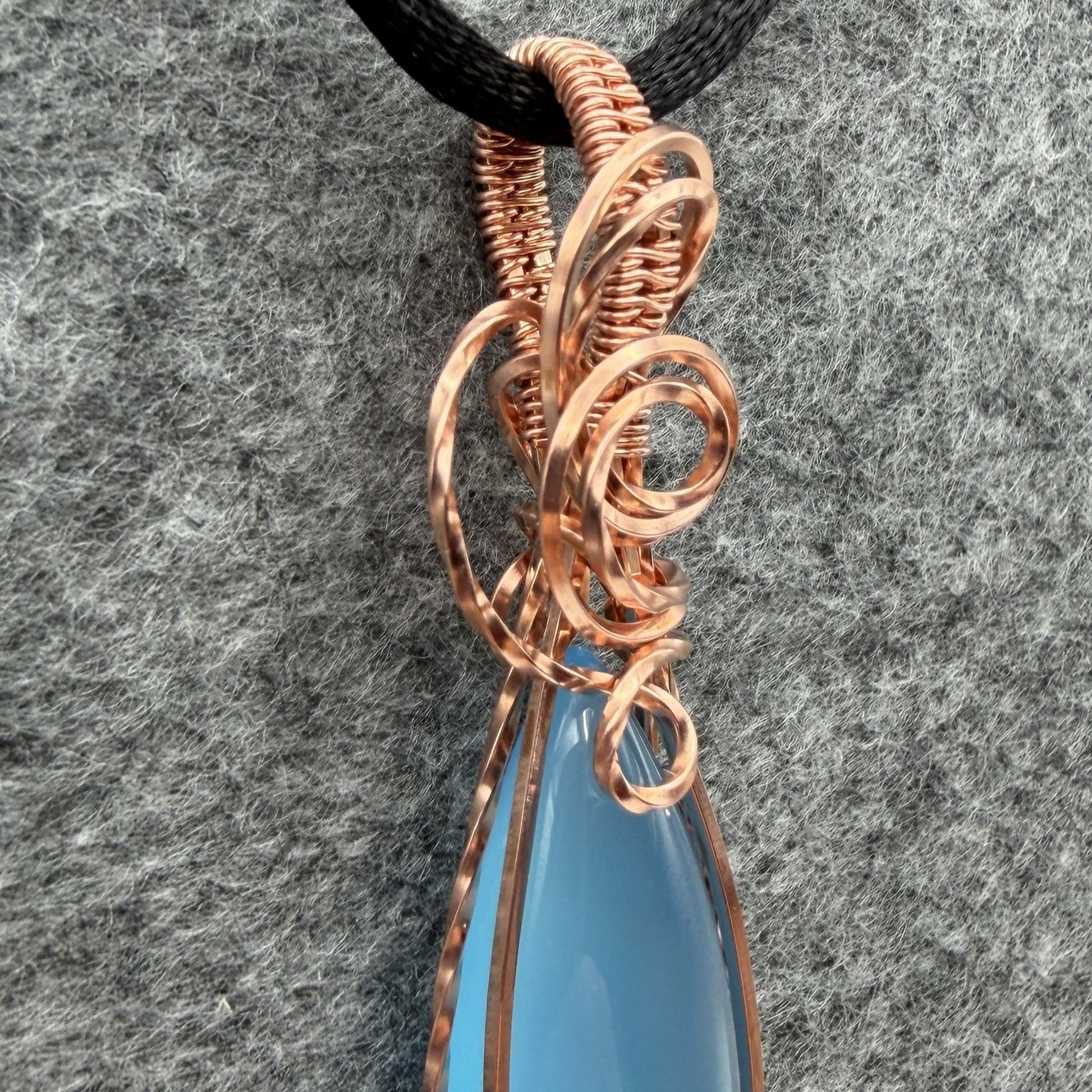 Detail view of a handmade pendant featuring a smooth, marquise-shaped light blue stone wrapped in textured copper wire, hanging from a black cord against a gray felt background.