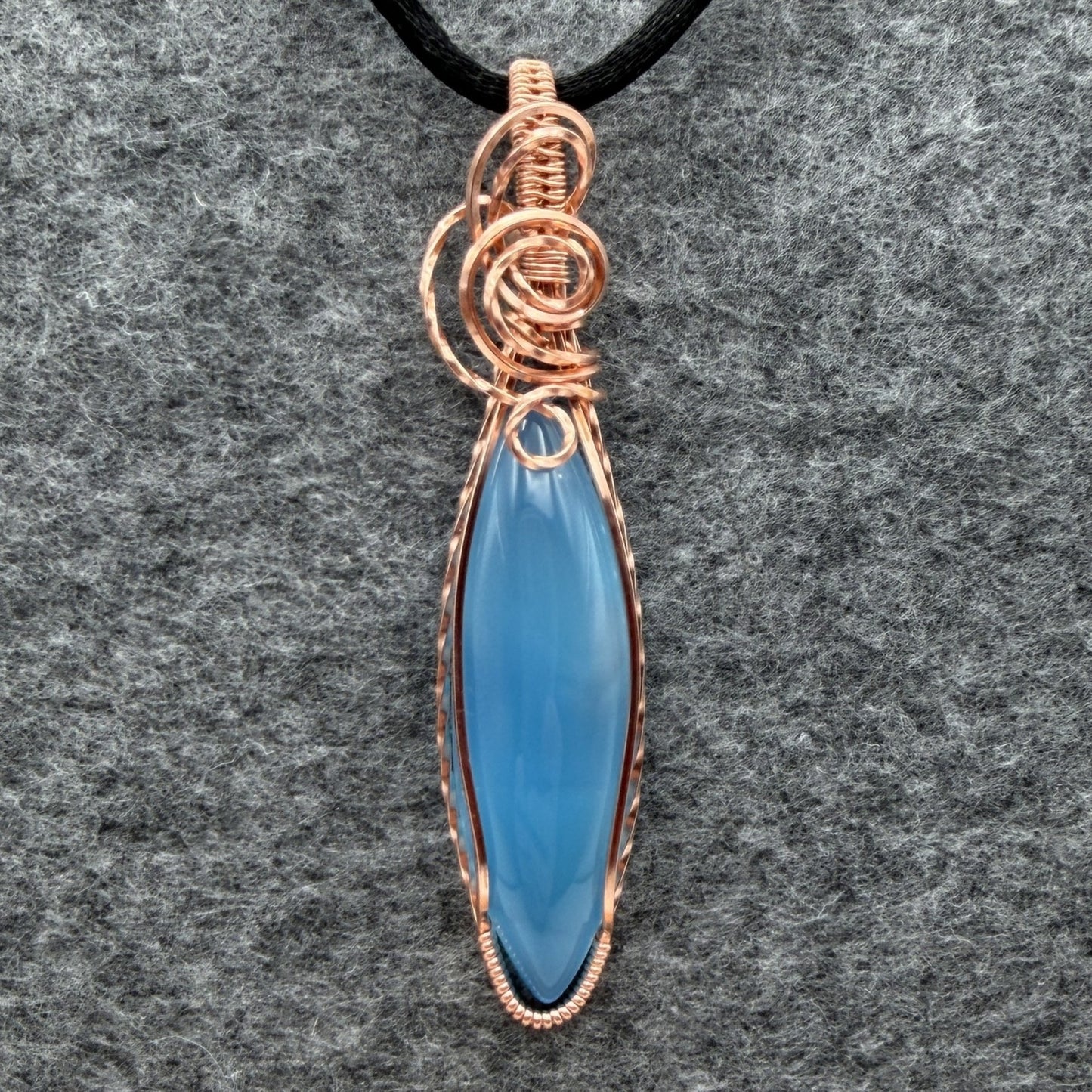 Front view of a handmade pendant featuring a smooth, marquise-shaped light blue stone wrapped in textured copper wire, hanging from a black cord against a gray felt background.