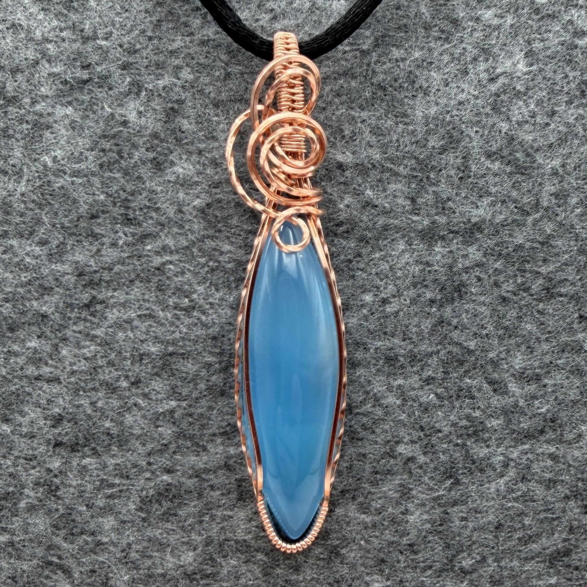 Front view of a handmade pendant featuring a smooth, marquise-shaped light blue stone wrapped in textured copper wire, hanging from a black cord against a gray felt background.