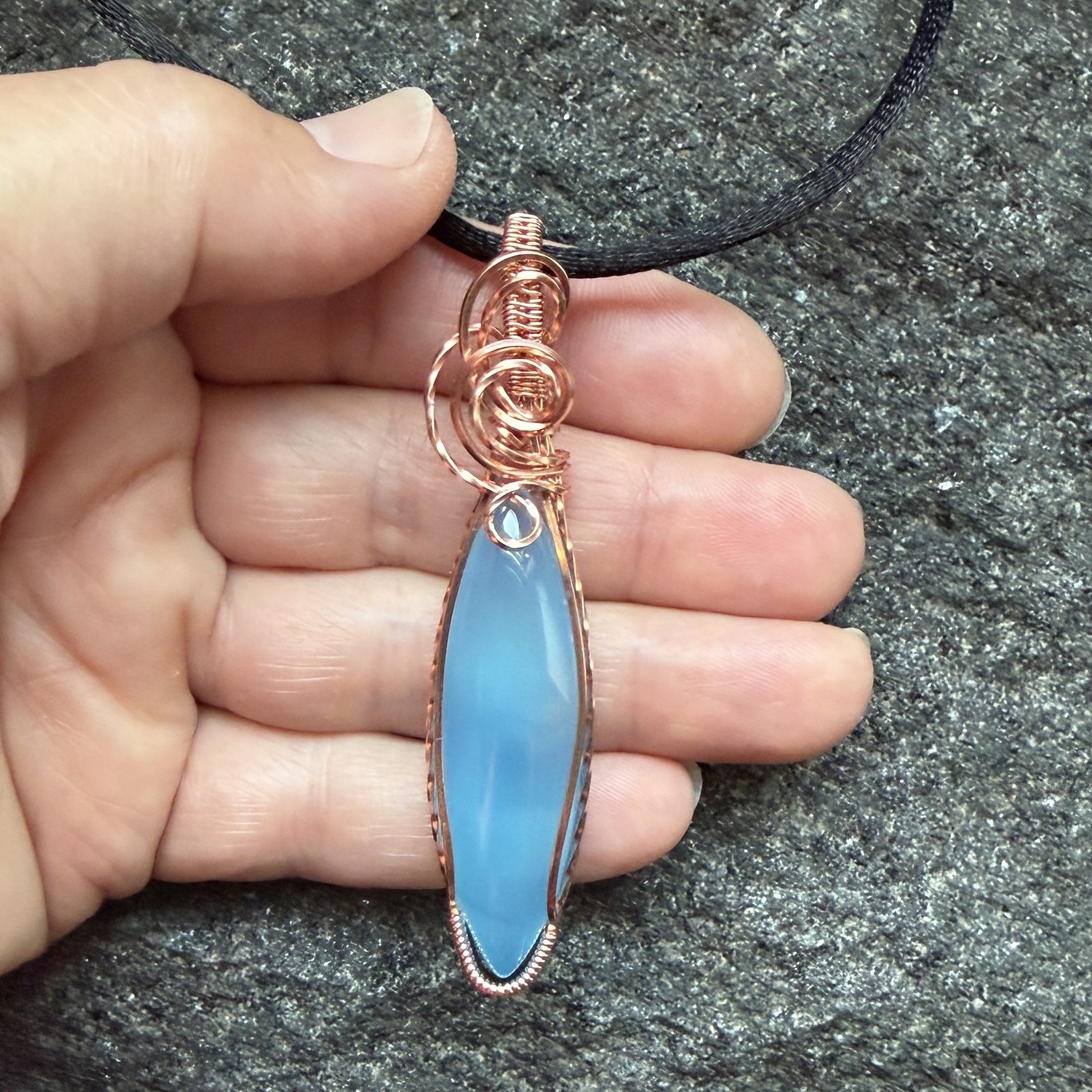 Front view of a handmade pendant featuring a smooth, marquise-shaped light blue stone wrapped in textured copper wire, hanging from a black cord and held in the artist's hand.