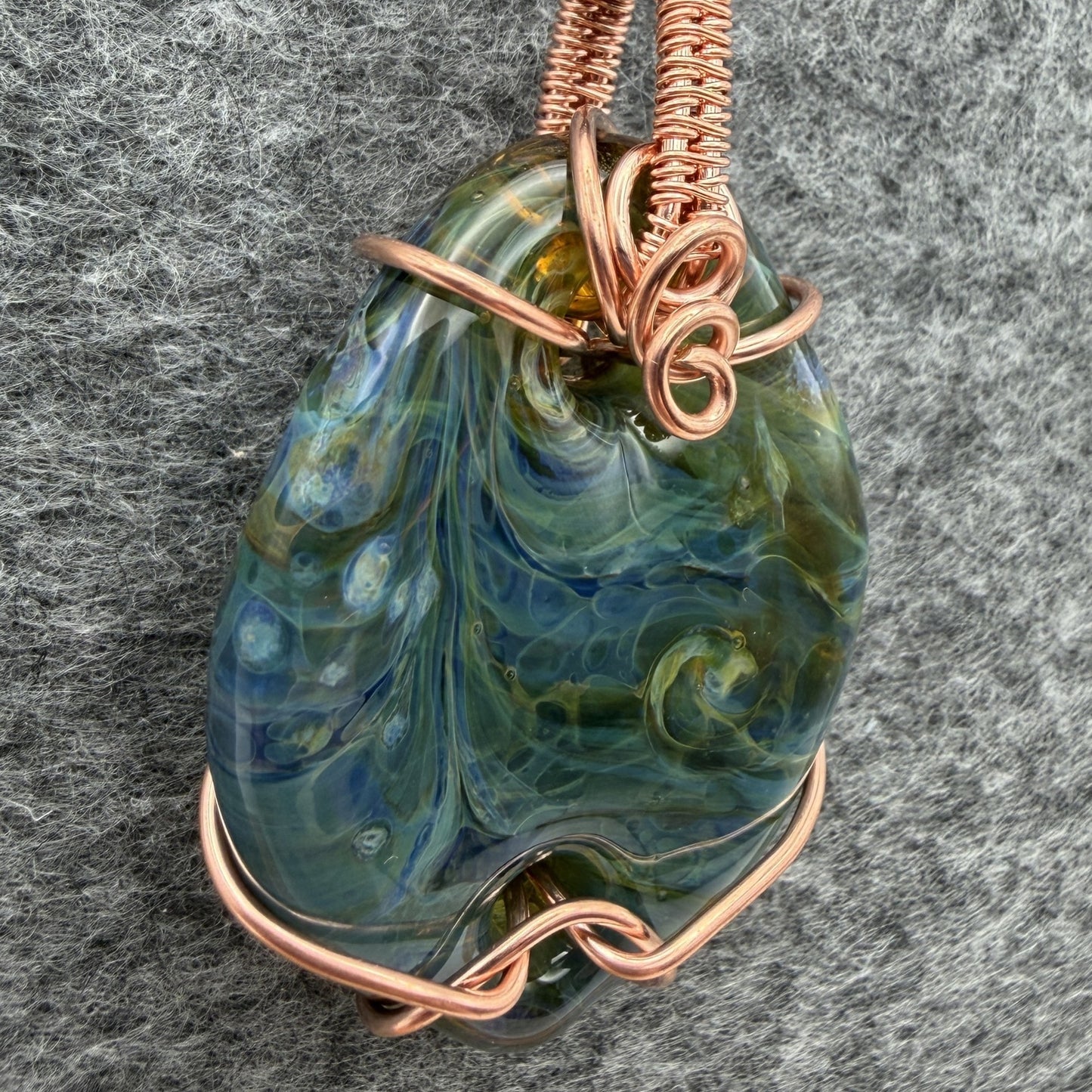 Detail view of side 2 of a handmade pendant necklace featuring a marbled blue and green glass stone wrapped in copper wire, hanging from a black cord against a textured gray background.
