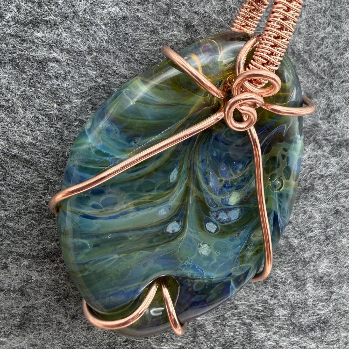 Detail view of side 1 of a handmade pendant necklace featuring a marbled blue and green glass stone wrapped in copper wire, hanging from a black cord against a textured gray background.